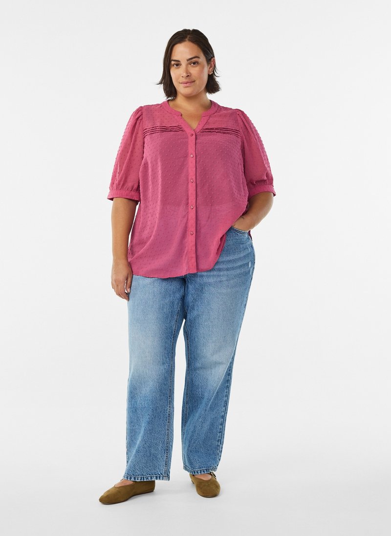 Mujer de pie con una mano en el bolsillo, vistiendo una blusa rosa, jeans azul claro y zapatos planos marrones sobre fondo blanco.