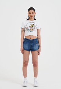 Witte cropped t-shirt met een bij en bloemenpatroon; gecombineerd met blauwe denim shorts. Model draagt witte sneakers en enkelsokken.