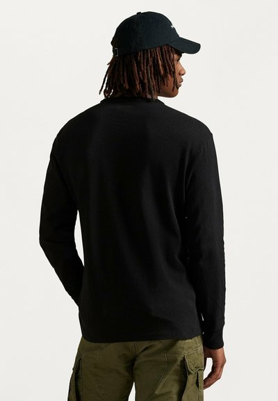 Maglietta nera a maniche lunghe con una texture liscia, abbinata a pantaloni cargo verde oliva e un cappellino nero, visibile da dietro.