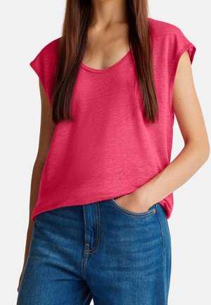 Mujer con una blusa sin mangas de color rosa brillante, metida ligeramente dentro de unos vaqueros azules, con una mano en el bolsillo.