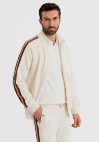 Homme portant un survêtement crème avec des rayures marron sur les manches et le pantalon, debout avec une main dans la poche, fond neutre.