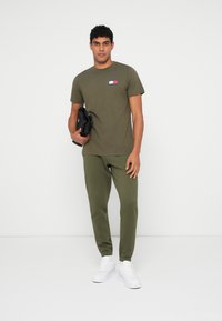 Ελαιόχρωμο μπλουζάκι με μικρό κόκκινο, λευκό και μπλε λογότυπο στο στήθος, συνδυασμένο με ταιριαστά παντελόνια φόρμας και λευκά αθλητικά παπούτσια.