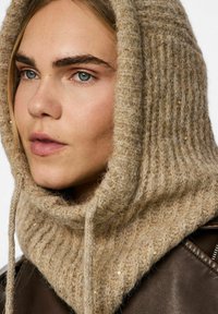 Beige knitted hood with a textured pattern, featuring subtle sequins, drapes casually around the neck. Paired with a brown leather jacket.