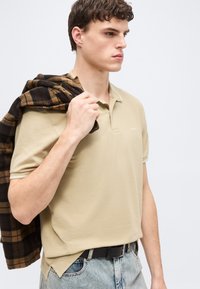 Jeune homme portant un polo beige et un jean clair, tenant une veste à carreaux marron et noire sur l'épaule, tourné vers la droite.