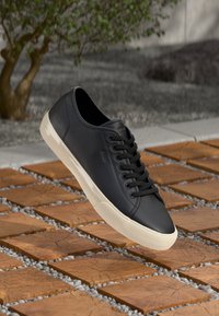 Black leather sneaker with flat laces, textured surface, and cream rubber sole. Elevated above patterned wood and gravel surface.