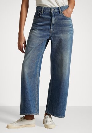 Vorderansicht einer Person, die ausgewaschene, gerade geschnittene blaue Jeans, weiße Sneaker und ein in die Hose gestecktes weißes Hemd trägt, stehend vor einem schlichten Hintergrund.
