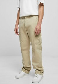 Pantalones cargo en beige, con grandes bolsillos laterales, presillas para el cinturón y corte relajado. Combinados con zapatillas blancas. Textura de tela suave.