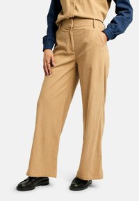 Nő bézs színű, széles szárú kordbársony nadrágban, tengerészkék hosszú ujjú ingben, fekete balerinacipőben, egyik kezével a zsebében, a másikkal a combján pihenve.