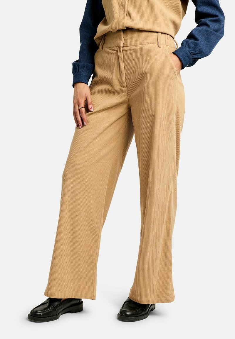 Nő bézs színű, széles szárú kordbársony nadrágban, tengerészkék hosszú ujjú ingben, fekete balerinacipőben, egyik kezével a zsebében, a másikkal a combján pihenve.