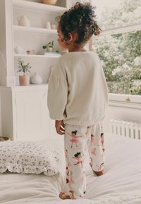 Hellbeige, oversized Sweatshirt und cremefarbene Schlafanzughosen mit Illustrationen von Balletttänzern, stehen auf einem Bett mit einer strukturierten weißen Decke.