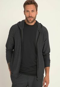 Felpa nera con zip e maniche lunghe, caratterizzata da una texture liscia e cappuccio con cordini, indossata sopra una t-shirt nera a girocollo.