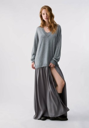 Le modèle porte un pull gris ample à col en V, une jupe longue fluide fendue, des bottes noires et un collier doré, sur un fond de studio uni.