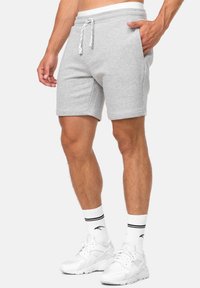 Shorts en coton gris clair avec une taille élastique, fermeture à cordon, poches latérales et coupe décontractée. Associés à des baskets blanches et des chaussettes rayées.