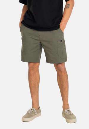 Mann trägt olivgrüne Cargo-Shorts, beige Sneakers und ein schwarzes T-Shirt, steht mit den Händen in den Taschen vor einem einfarbigen Hintergrund.