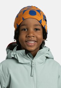 Orange beanie with blue patterns, made of knit material. Worn with a light green, zippered, hooded jacket.