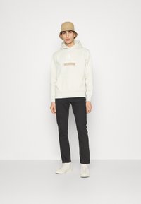 Felpa beige chiaro con tasca frontale, logo bianco, abbinata a pantaloni neri slim-fit e sneaker bianche; il modello indossa un cappello a secchiello color sabbia.