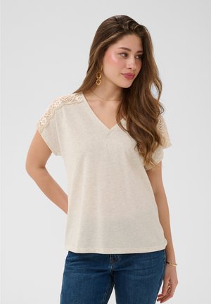 Femme aux longs cheveux bruns portant un haut beige à manches courtes avec des détails en dentelle aux épaules et un jean bleu, regardant légèrement sur le côté.