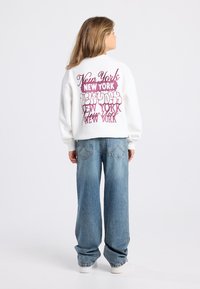 Jeune personne avec de longs cheveux portant un sweat-shirt blanc avec plusieurs textes "New York" dans le dos, un jean bleu et des baskets blanches, se tenant de profil.