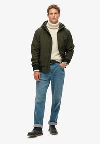 Grøn nylonjakke med hætte, lys beige turtleneck, lyseblå lige jeans og sorte snørestøvler, stående på en hvid baggrund.