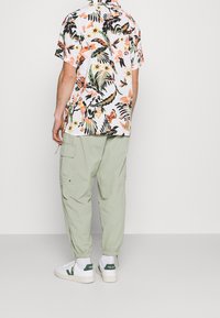 Chemise à manches courtes florale avec des plantes vibrantes et des papillons, assortie à un pantalon cargo vert clair et des baskets blanches avec des accents verts.
