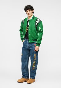 Πράσινο varsity μπουφάν με λευκές λεπτομέρειες και γράμματα, μαύρο T-shirt, μπλε τζιν με γραφικά μοτίβα και καφέ μπότες.