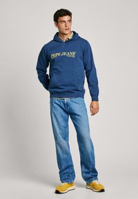 Temno modra kapuca z rumenim logotipom, kenguru žepom in nastavljivim vezalko za kapuco. Ujemajoče se z svetlo modrimi kavbojkami in rumenimi supergami.