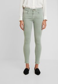 Jeans ajustados verdes de mezclilla. Presentan bolsillos frontales, cierre de botón y una textura suave. Combinados con mocasines negros.