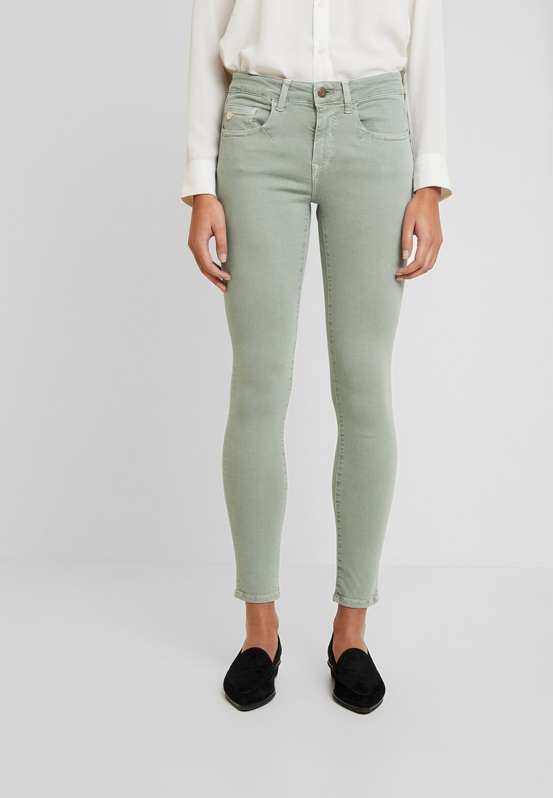 Jeans ajustados verdes de mezclilla. Presentan bolsillos frontales, cierre de botón y una textura suave. Combinados con mocasines negros.