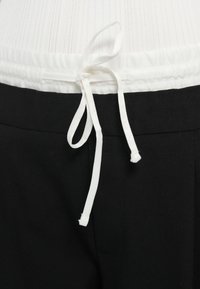 Pantalones negros con una pretina elástica blanca y detalle de lazo. La tela tiene un acabado texturizado e incluye bolsillos laterales.