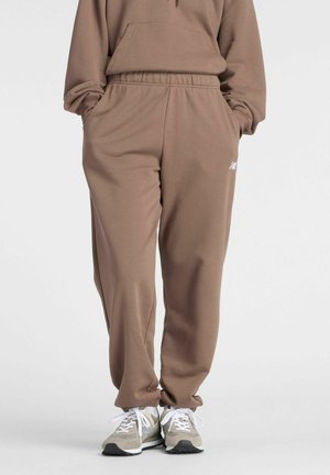 Personne portant un pantalon de survêtement marron à coupe décontractée et un sweat à capuche assorti avec des baskets blanches et beige, debout les mains dans les poches.