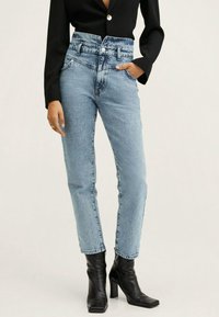 Hochtaillierte blaue Jeans aus Denim mit leichter Textur, mit Knopfverschluss, Fronttaschen und schmal zulaufendem Bein. Getragen mit schwarzen Absatzstiefeln.