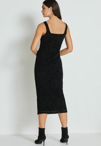 Sort midi-kjole med et tekstureret mønster, firkantet udskæring og brede stropper. Sorte ankelstøvler komplementerer outfittet og fremhæver det strømlinede design.