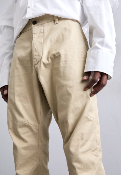 Pantaloni in cotone beige con texture sottile, dotati di tasca anteriore, passanti per cintura e dettagli cuciti sui lati. Abbinati a una camicia bianca.