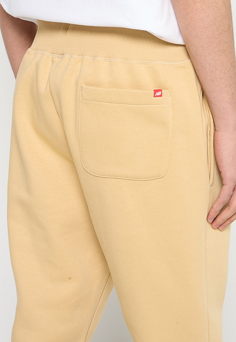 Pantalones sudadera color beige claro con cintura acanalada y un bolsillo trasero. Detalle de etiqueta roja en el bolsillo. Textura de tela suave y lisa.
