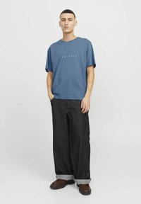 Blaues T-Shirt mit kurzen Ärmeln und weißem Aufdruck, kombiniert mit dunkelblauen, weiten Jeans. Die Hose hat umgeschlagene Bündchen und kontrastierende gelbe Nähte.
