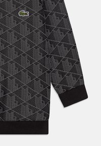 Czarny i szary sweter z nadrukiem o geometrycznych wzorach, z prążkowanymi mankietami oraz małym zielonym logo Lacoste na klatce piersiowej.