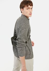 Grauer Strickpullover mit hohem Kragen, getragen mit einer schwarzen Umhängetasche mit Reißverschlüssen und kontrastierenden Trägern, vor einem weißen Hintergrund.