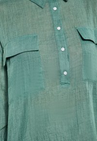 Camicia verde a bottoni realizzata in tessuto leggero, con finitura testurizzata, due tasche sul petto e bottoni bianchi.