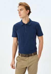 Marineblauwe poloshirt met een ritssluiting bij de kraag, korte mouwen, gemaakt van een soepele katoenmix. Gecombineerd met lichtbeige broeken en een bruine riem.