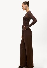 Haut marron transparent à manches longues avec des clous argentés, associé à un pantalon marron taille haute à jambes larges. Les manches allongées sont une caractéristique notable.