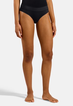 Femme debout pieds nus portant un sous-vêtement noir taille haute et sans coutures sur un fond clair uni.