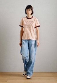 Απαλή ροζ βαμβακερή μπλούζα με μπορντό λεπτομέρειες και γραφικό μπροστά, συνδυασμένη με φαρδιά, ξεθωριασμένα τζιν και λευκά αθλητικά παπούτσια πάνω σε ξύλινο πάτωμα.