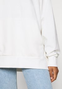 Felpa bianca oversize realizzata in morbido tessuto, con tasca anteriore, polsini e orlo a costine. Indossata con jeans in denim azzurro chiaro.