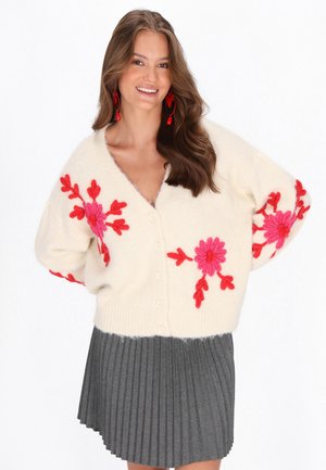 Cardigan crema con vivaci ricami floreali rosa, caratterizzato da scollatura a V e chiusura con bottoni. Abbinato a una gonna grigia a pieghe.