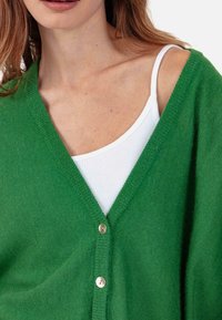 Cardigan tricoté vert avec encolure en V et trois boutons argentés. En dessous, un débardeur blanc. Le tissu semble doux et texturé.