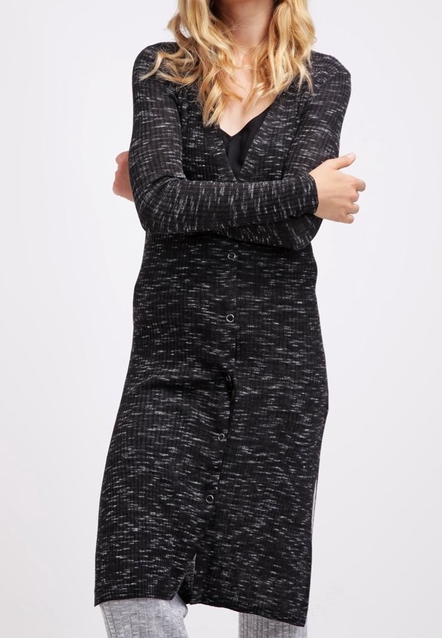 Femme bras croisés portant un long cardigan noir et gris moucheté par-dessus un haut noir et un pantalon gris clair sur un fond uni.