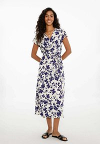 Kvetinové zavinovacie šaty v krémovej farbe s námorníkovým kvetinovým vzorom, krátke rukávy, po kolená, s previazaným pásom, skombinované s čiernymi sandálmi. Hladká textúra látky.