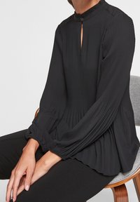 Svart plisserad blus med hög krage och knappstängning, med långa puffärmar och avslappnad passform, kombinerad med svarta byxor.