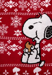 Un tessuto lavorato a maglia rosso presenta un cane dei cartoni animati bianco e nero che tiene un biscotto marrone. Lo sfondo include motivi di fiocchi di neve bianchi sparsi.