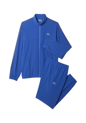 Giacca a zip blu e pantaloni da tuta con vita elastica coordinati, entrambi con un piccolo logo di coccodrillo verde.